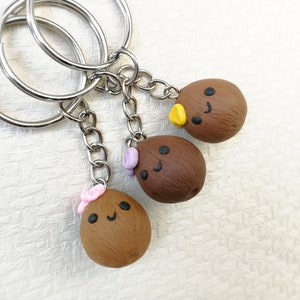 May include: A keychain with three brown coconut-shaped charms. Each charm has a different facial expression and a small decorative detail. The charms are attached to a silver key ring.