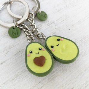 May include: Two avocado-shaped keychains with smiling faces, pink cheeks, and brown heart-shaped pits. Each keychain has a silver chain and a green circular charm with a letter. The avocados are green and yellow.