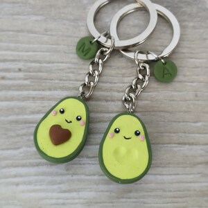 May include: Two avocado-shaped keychains with smiling faces and silver key rings. One avocado has a brown heart, the other has a pit. Each keychain has a green charm with a letter. The keychains are on a light wood surface.