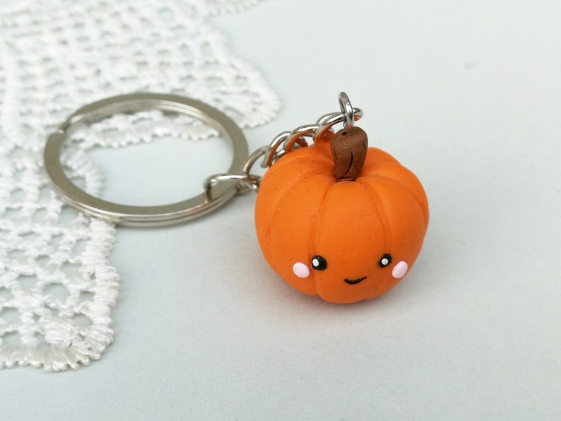 May include: A small orange pumpkin keychain with a brown stem and a cute smiling face. The pumpkin has pink cheeks and is attached to a silver key ring.