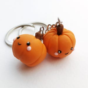 May include: Two orange pumpkin-shaped keychains with smiling faces and pink cheeks. The keychains are attached to a silver key ring.