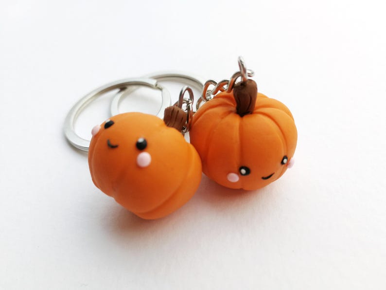 May include: Two orange pumpkin-shaped keychains with smiling faces and pink cheeks. The keychains are attached to a silver key ring.