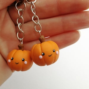 May include: Two orange pumpkin keychains with happy faces and pink cheeks. The keychains are attached to silver chains.