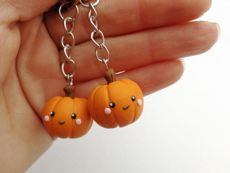 May include: Two orange pumpkin keychains with happy faces and pink cheeks. The keychains are attached to silver chains.