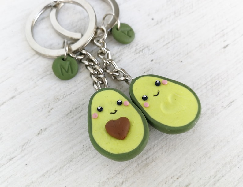 May include: Two avocado-shaped keychains with smiling faces, pink cheeks, and brown heart-shaped pits. Each keychain has a silver chain and a green circular charm with a letter. The avocados are green and yellow.