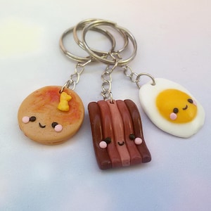 Puede incluir: Tres llaveros adorables con tem&aacute;tica de comida: un panqueque con una cara sonriente y una sola chispa de chocolate, una tira de tocino con una cara sonriente y un huevo frito con una cara sonriente.