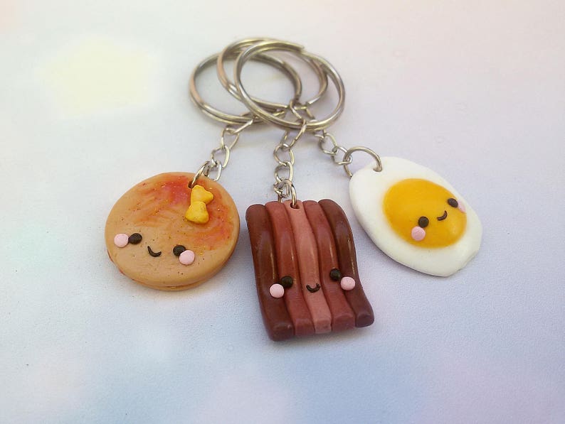 Puede incluir: Tres llaveros adorables con tem&aacute;tica de comida: un panqueque con una cara sonriente y una sola chispa de chocolate, una tira de tocino con una cara sonriente y un huevo frito con una cara sonriente.