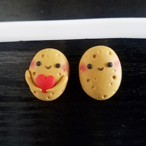 Puede incluir: Dos figuras caprichosas en forma de patata. Una sostiene un corazón rojo, la otra tiene mejillas rosadas y una cara sonriente. Ambas tienen pequeños ojos negros y son de color marrón claro con pequeñas hoyuelos. Las figuras están sobre una superficie oscura.