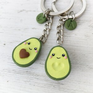 May include: Two avocado-shaped keychains with smiling faces and heart details. The keychains are light green with a dark green border and a silver chain. One has a brown heart, the other a heart-shaped indentation. Each has a letter charm.