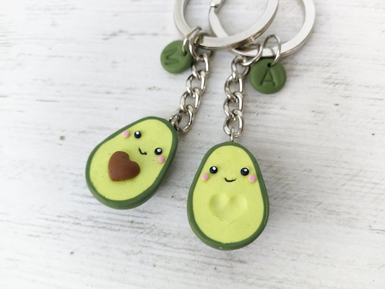 May include: Two avocado-shaped keychains with smiling faces and heart details. The keychains are light green with a dark green border and a silver chain. One has a brown heart, the other a heart-shaped indentation. Each has a letter charm.