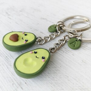 May include: Two avocado-shaped keychains with smiling faces. The keychains are green and light green with a brown pit. Each keychain has a silver chain and a silver keyring. One keychain has a green charm with the letter "A".