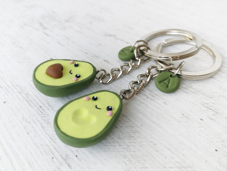 May include: Two avocado-shaped keychains with smiling faces. The keychains are green and light green with a brown pit. Each keychain has a silver chain and a silver keyring. One keychain has a green charm with the letter "A".