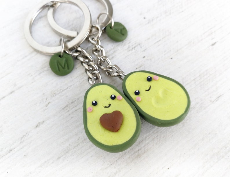 May include: Two avocado-shaped keychains with smiling faces, pink cheeks, and brown avocado pit details. Each keychain has a silver chain and ring, with a green charm bearing a letter. The avocados are light green with a darker green border.