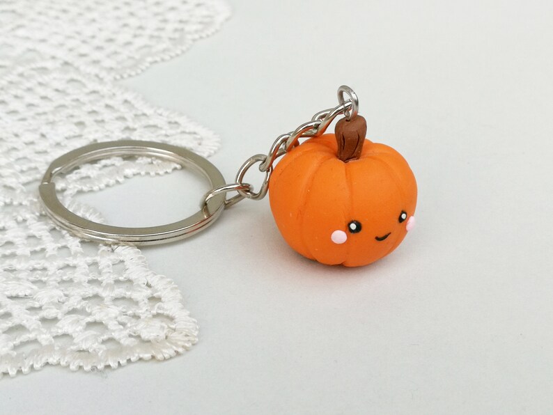May include: A small orange pumpkin keychain with a brown stem and a cute cartoon face. The pumpkin has pink cheeks and a small smile.