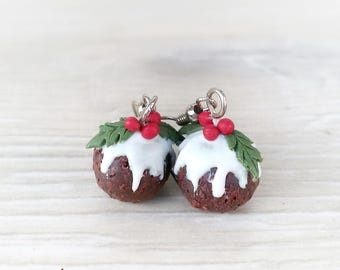 May include: A pair of Christmas pudding earrings. Each earring is a small, round, dark brown pudding with white icing drizzled over the top. Green holly leaves and red berries adorn the top of each pudding. The earrings hang from silver-colored hooks.