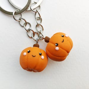 May include: Two orange pumpkin-shaped keychains with happy faces. The keychains are attached to a silver chain.