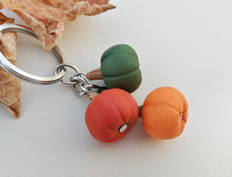 Puede incluir: Un llavero de plata con tres peque&ntilde;as calabazas decorativas. Una calabaza es verde, una es naranja y una es roja.