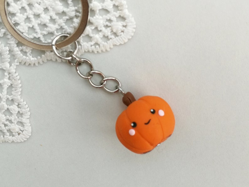 May include: A cute orange pumpkin keychain with a smiling face and rosy cheeks. The pumpkin is attached to a silver key ring.