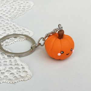 May include: Orange pumpkin keychain with a cute smiling face and pink cheeks. The pumpkin is attached to a silver key ring.