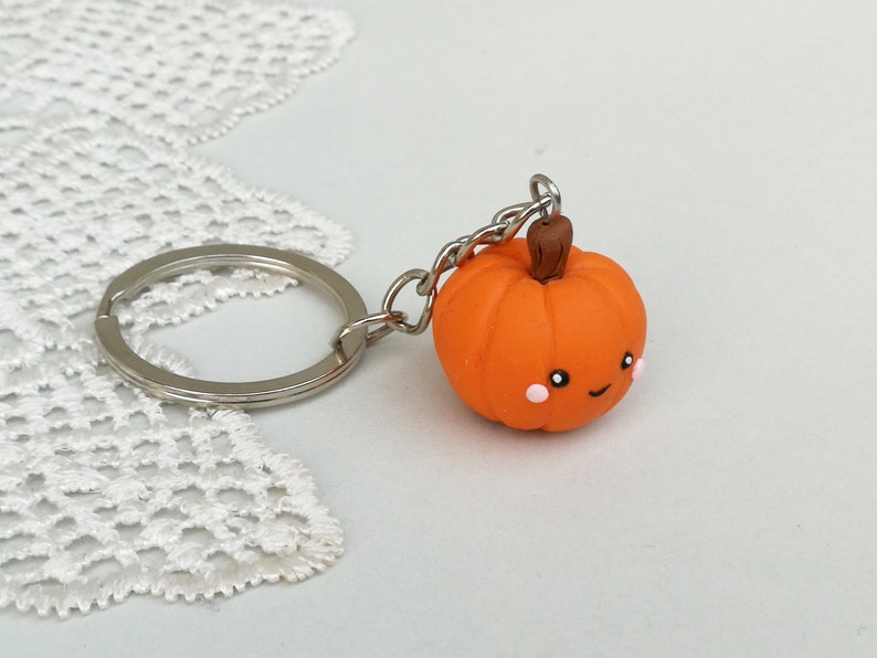 May include: Orange pumpkin keychain with a cute smiling face and pink cheeks. The pumpkin is attached to a silver key ring.