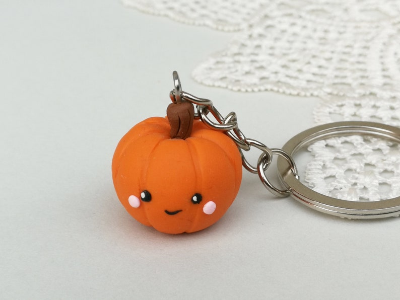 May include: A small orange pumpkin keychain with a brown stem and a cute smiling face. The pumpkin has pink cheeks and black eyes. The keychain is attached to a silver key ring.