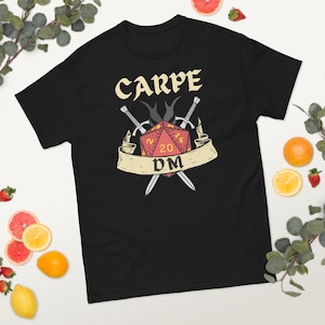 Puede incluir: Camiseta negra con un dado rojo y amarillo de 20 caras, dos espadas cruzadas y el texto "CARPE DM" en una pancarta.