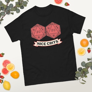 以下が含まれることがあります： 黒いTシャツ。ピンク色の20面ダイスが2つ描かれ、数字が見えます。ダイスの下には、赤い縁取りのある白い文字で「NICE CRITS」と書かれたバナーがあります。クラシックフィットです。