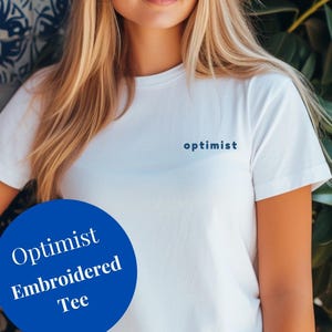 Op de afbeelding: Wit T-shirt met het woord "optimist" in het blauw geborduurd op de voorkant.