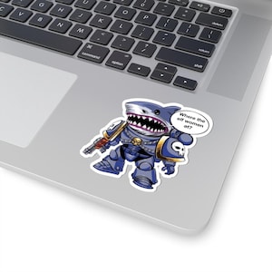 Peut inclure: Un autocollant représentant un requin de dessin animé en armure bleue, tenant une arme. Le requin a un large sourire et une bulle de dialogue qui dit : "Where the elf women at ?" L'autocollant est sur un ordinateur portable.