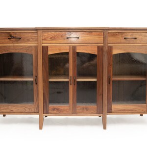 May include: A three-section wooden cabinet with glass doors and shelves. Each section has a drawer with a black metal pull. The cabinet is made of dark wood with a natural finish.