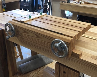 May include: A wooden workbench with a vise and a wooden top. The vise has two metal wheels and a wooden handle. The workbench is made of light wood and has a natural finish.