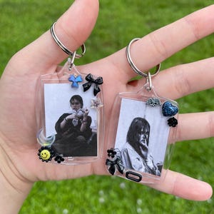 May include: Two clear plastic keychains with photos inside. The keychains are decorated with small charms, including a butterfly, a heart, a flower, and a crescent moon.