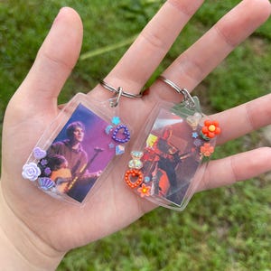 May include: Two clear plastic keychains with photos inside. The keychain on the left has a photo of a man holding a guitar and is decorated with purple, blue, and white embellishments. The keychain on the right has a photo of a band performing and is decorated with orange, red, and white embellishments.