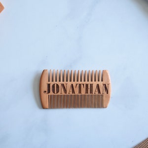 Może przedstawiać: Drewniany grzebień z wygrawerowanym imieniem "JONATHAN". Grzebień ma zaokrąglony kształt i jest wykonany z jasnobrązowego drewna.