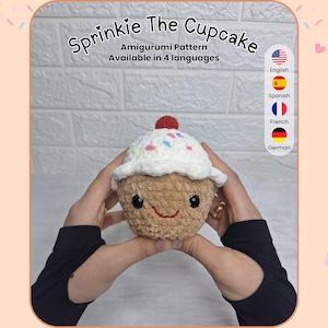 Könnte beinhalten: Ein gehäkelter Cupcake-Amigurumi mit einem lächelnden Gesicht, weißem Zuckerguss und bunten Streuseln. Der Cupcake hat eine rote Kirsche oben drauf. Der Text "Sprinkie The Cupcake" und "Amigurumi Pattern Available in 4 languages" sind sichtbar.