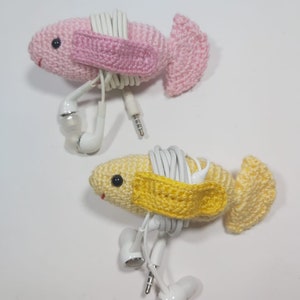 May include: Two crocheted fish, one pink and one yellow, with white headphone cords wrapped around them. The fish have a small crocheted strap across their bodies to hold the cords in place.