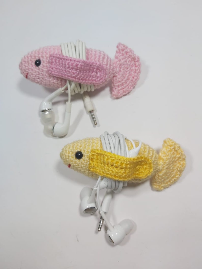May include: Two crocheted fish, one pink and one yellow, with white headphone cords wrapped around them. The fish have a small crocheted strap across their bodies to hold the cords in place.