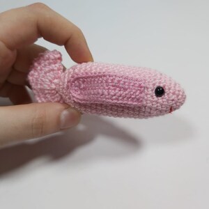 May include: A pink crocheted fish toy with black eyes. The fish is small and is being held in a hand.