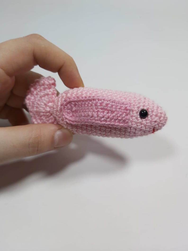 May include: A pink crocheted fish toy with black eyes. The fish is small and is being held in a hand.