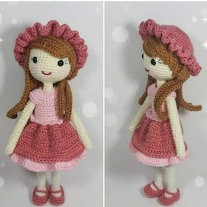 Puede incluir: Una muñeca de crochet con cabello castaño, un vestido rosa y un sombrero rosa. La muñeca está de pie sobre un fondo blanco.