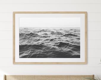 May include: A wooden bench with a white and grey checked cushion and two throw pillows. The bench is in front of a white wall with a framed black and white photograph of ocean waves. A potted plant is on the right side of the bench.