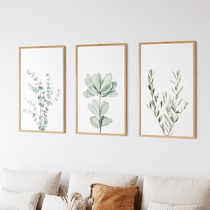 May include: Three framed botanical prints with watercolor illustrations of green leaves on a white background. The prints are hung on a white wall above a light brown couch with white cushions and a brown throw pillow.