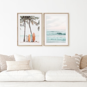 May include: Two framed prints with a beach theme. The first print shows a row of colorful surfboards leaning against a palm tree on a sandy beach. The second print shows a turquoise ocean with white waves crashing on the shore.