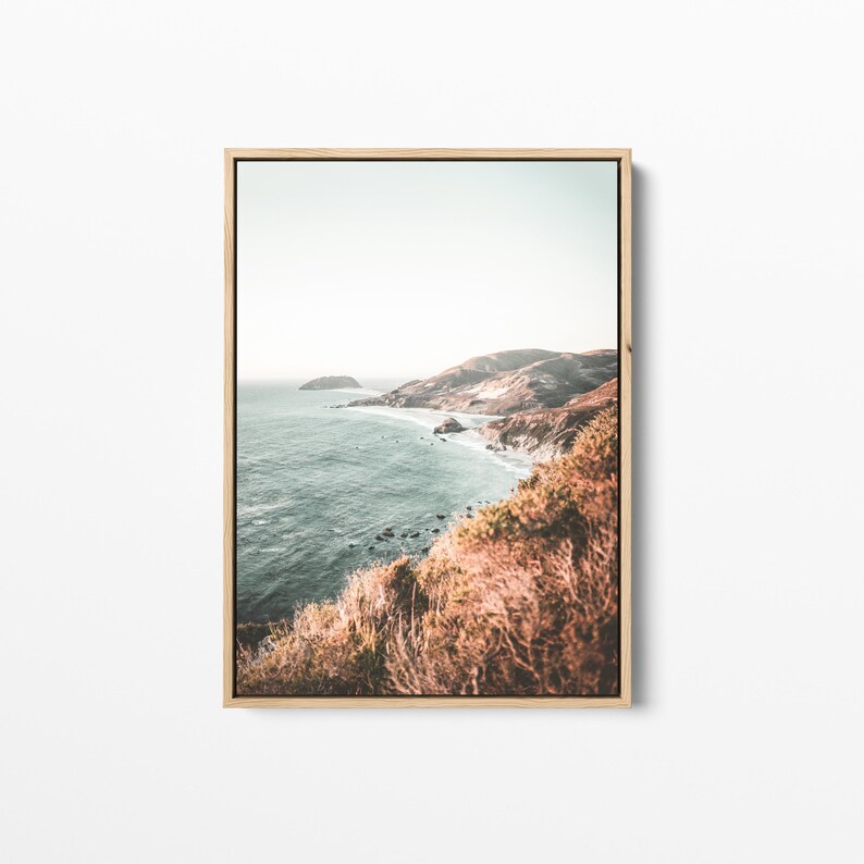 May include: A framed photograph of a coastal landscape with a view of a rocky coastline and the ocean. The photo is taken from a high vantage point, looking down at the water and the shore. The colors in the photo are muted and natural, with shades of blue, green, and brown.