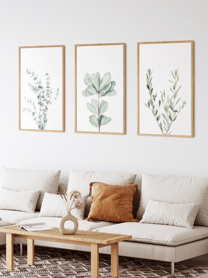 May include: Three framed botanical prints with watercolor illustrations of green leaves on a white background. The prints are hung on a white wall above a light brown couch with white cushions and a brown throw pillow.