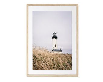 May include: A framed print featuring a coastal scene with a white lighthouse under a cloudy sky. The lighthouse has a black top and sits atop a grassy hill. The print is framed in a light wood frame.