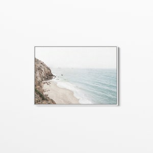 May include: A photograph of a rocky cliff overlooking a sandy beach and the ocean. The water is a light blue color and the sky is a hazy white.
