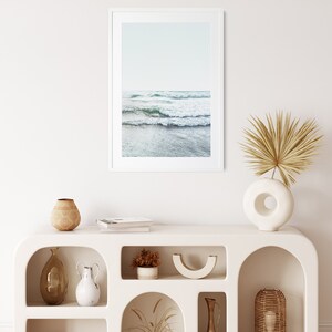 May include: White shelf unit with arched openings, featuring various decorative items, including a framed photograph of ocean waves, a vase with dried palm leaves, a wooden bowl, and a ceramic vase.