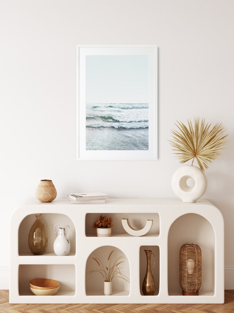 May include: White shelf unit with arched openings, featuring various decorative items, including a framed photograph of ocean waves, a vase with dried palm leaves, a wooden bowl, and a ceramic vase.