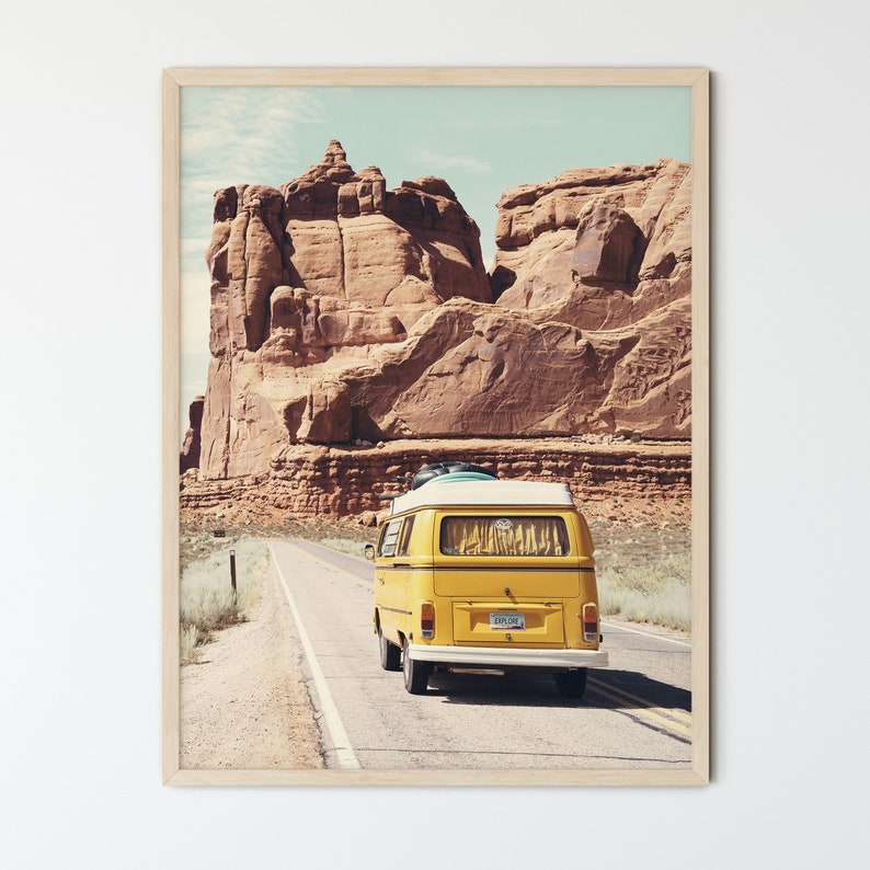 May include: A yellow vintage van drives down a paved road in a desert landscape. The van has a surfboard strapped to the roof and a license plate that reads "EXPLORE".